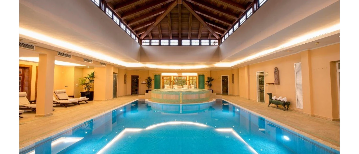 Rectangular swimming pool with LED lighting and a raised whirlpool section, in a room with a high wood-beamed ceiling, white columns and loungers