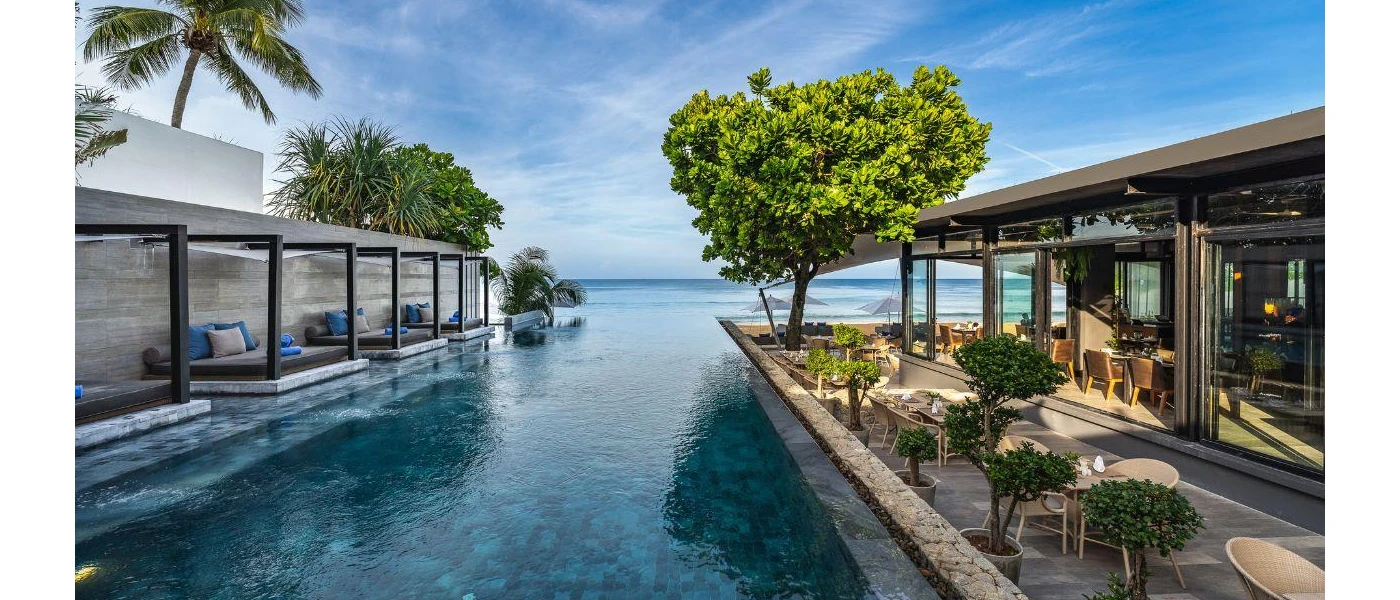 Infinity pool with ocean views and trees, lined on one side by grey-cushioned daybeds and the other by a leafy terrace with rattan chairs and tables