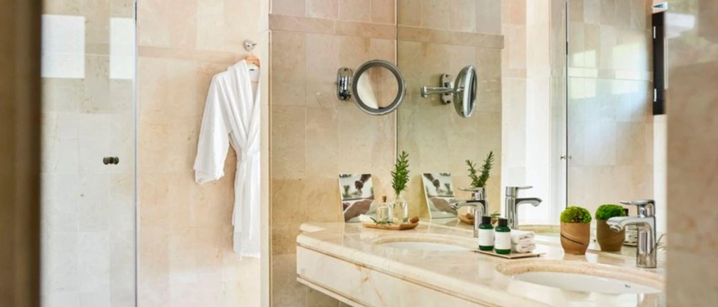 Cream marble bathroom with a white robe hanging on the wall, a vanity unit with two sinks and potted green plants, a shower and a large mirror, plus a magnifying mirror coming out of the wall