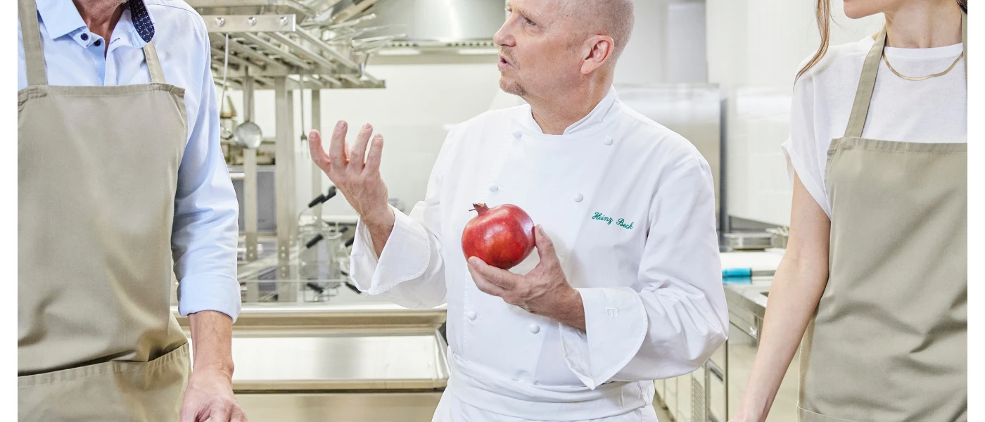Chef in whites holding a red vegetable gesturing with two people in aprons next to him