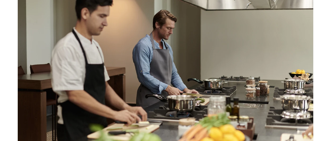 Two men in aprons stand at hobs while enjoying a cooking class, with fresh ingredients around them