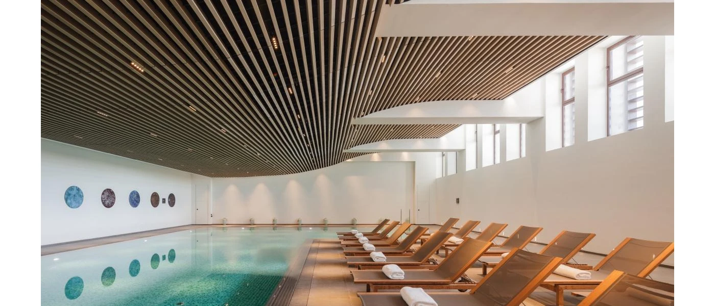 Indoor pool with a neighbouring terrace lined with loungers, each with a rolled white towel on top
