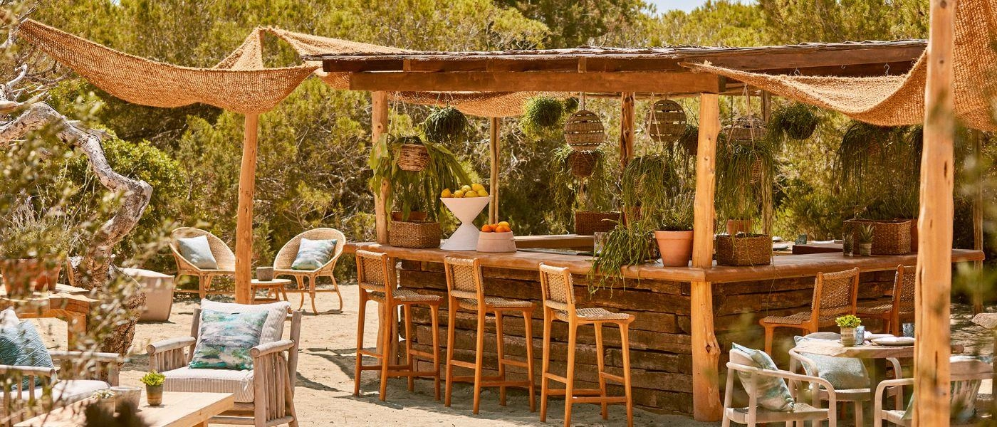 Boho-style bar with wooden stilts, rattan canopies and surrounding pine trees, with wooden seating and blue cushions