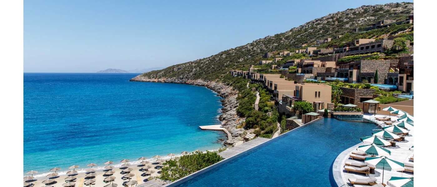 Low-rise villas, an infinity-edge pool and terrace with white-cushioned loungers and umbrellas, on a hillside overlooking a sandy beach furnished with loungers and umbrellas and overlooking a turquoise sea