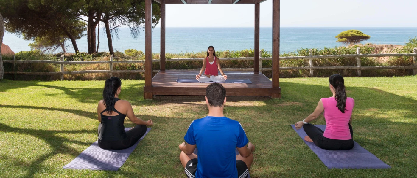 Three people in active wear practice yoga on mats in an ocean-facing garden, as an instructor leads from a wooden cabana at the front of the class