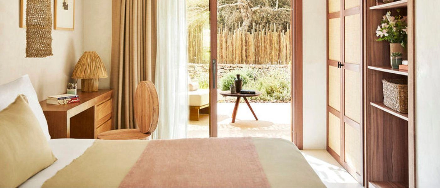 Contemporary boho-style hotel bedroom with neutral soft furnishings, wooden furniture and a terrace fringed by greenery