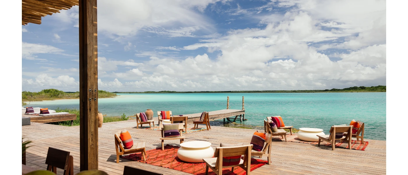 Wooden deck on the waterfront with wooden chairs with orange cushions, orange blankets and bean bags overlooking the lagoon
