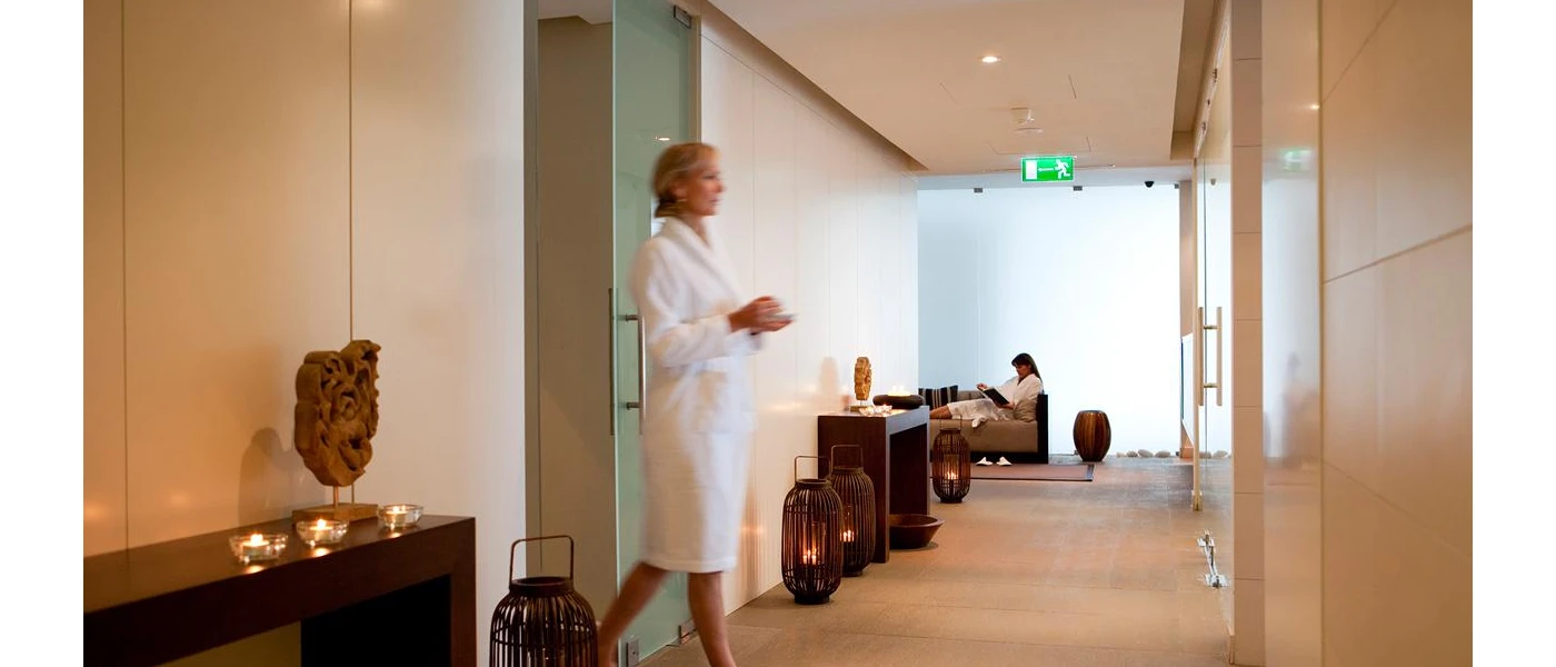 Woman in a white robe walks through a corridor with cream walls and flooring, ceiling spotlights and dark-wood units with glass tealight candle holders and wooden sculptures