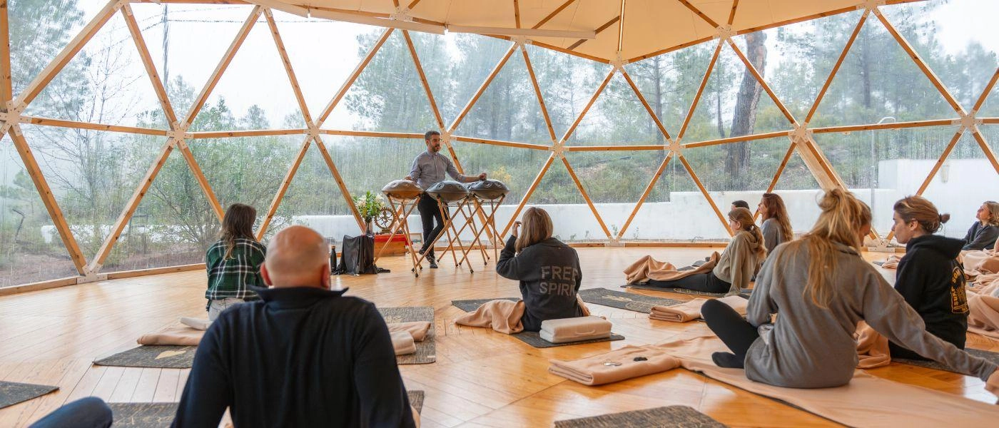 Group sits in a circle in a sheer domed building, while a man at front is playing sound healing bowls