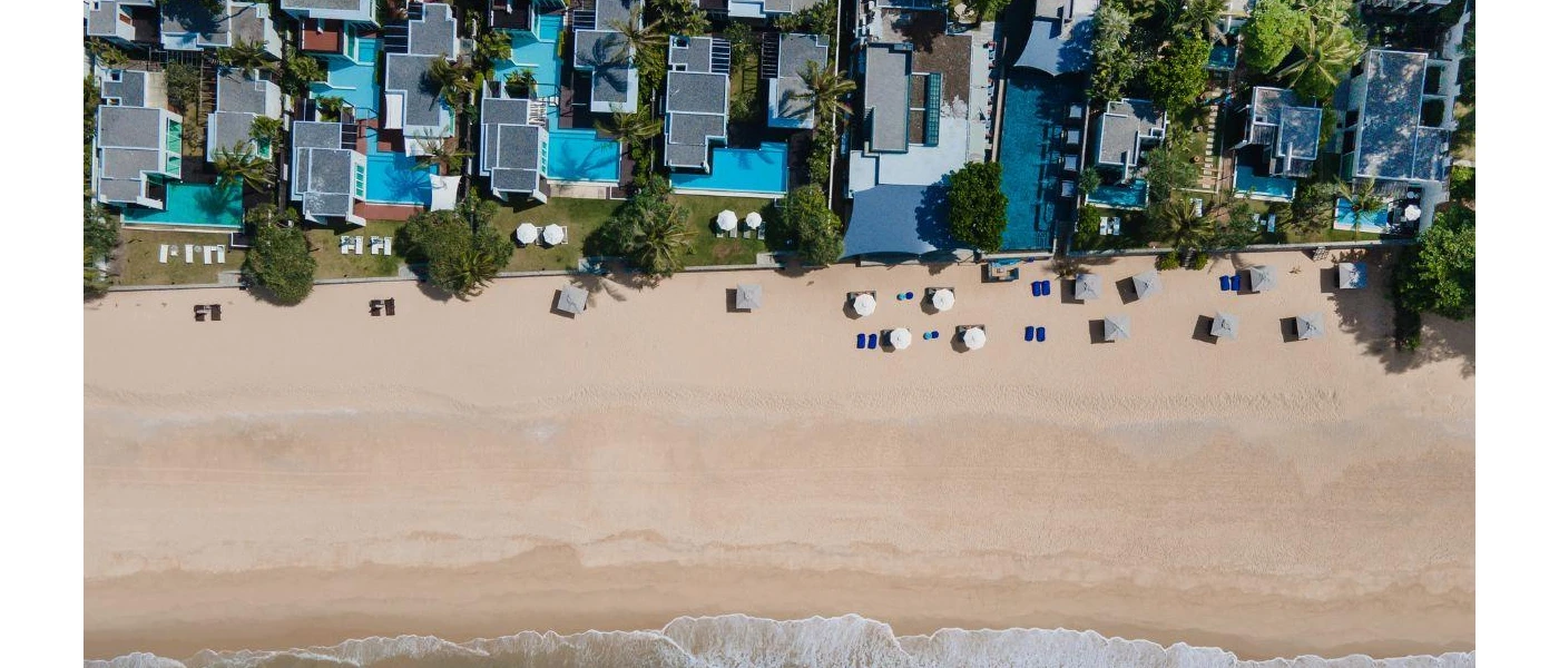 Aerial view of a sandy beach, white villas with private pools and surrounding greenery