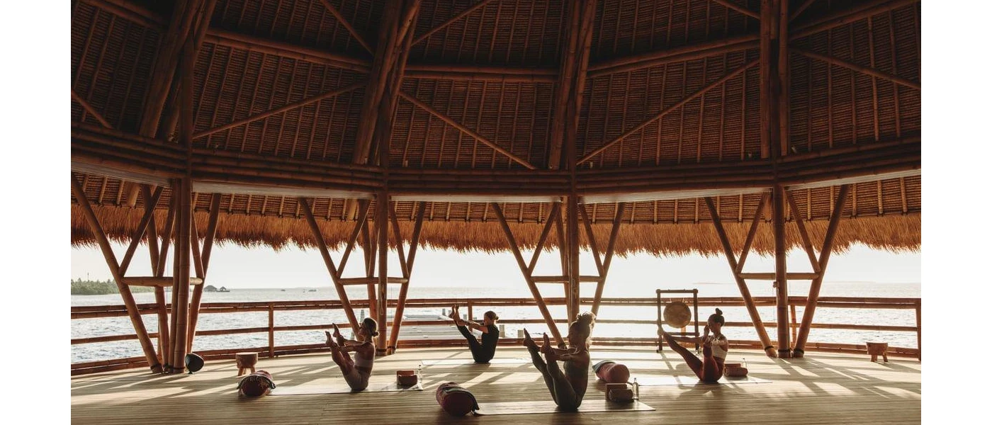 Group of flexible woman holding their ankles while stretching their legs upwards, on mats in an open-air thatched-roof pavilion