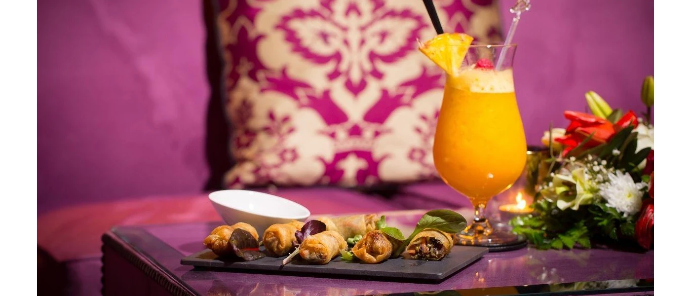Purple patterned cushion next to an orange cocktail and appetisers on a black tray 