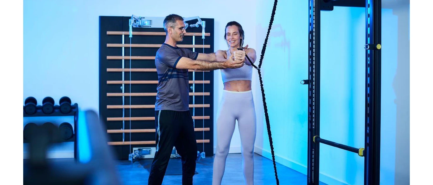 Woman in light leggings and a lilac gym top stretches a rope in a gym, supported by a personal trainer