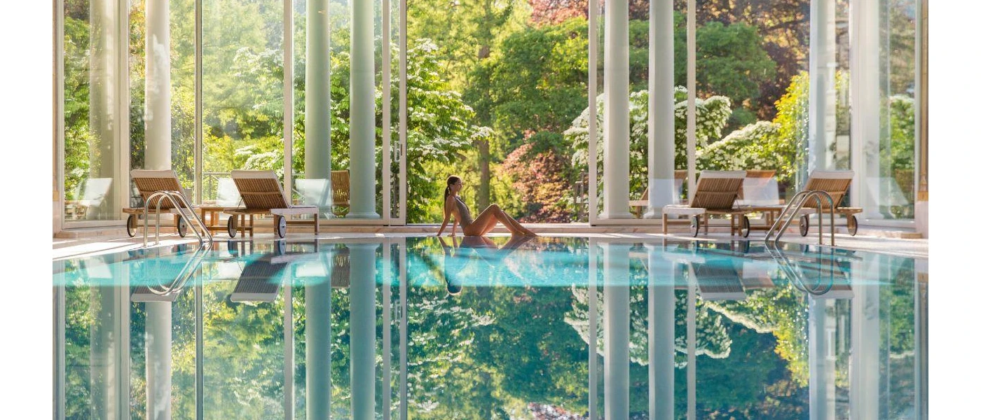 Toned woman in a swimsuit sits at the edge of an indoor pool surrounded by white pillars and wooden, white-cushioned loungers, with floor-to-ceiling glass in the background showcasing the gardens beyond