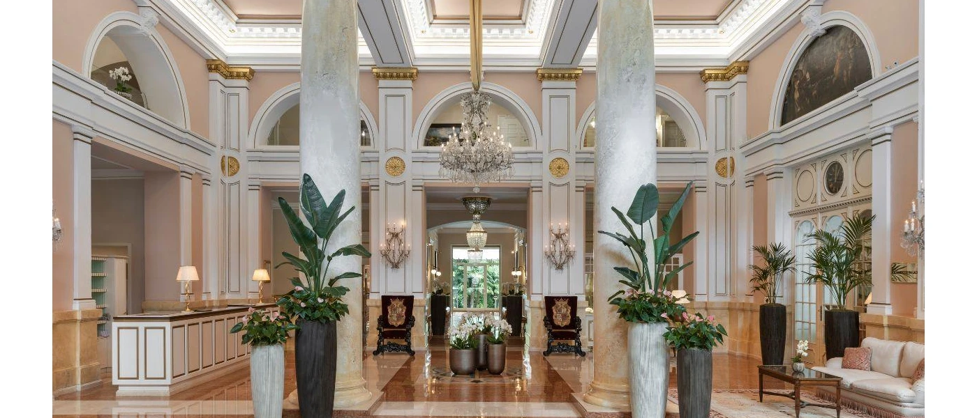 High ceilinged lobby area with ornate rugs and armchairs, gold accents, glittering chandelier-stye wall lights and grand white pillars
