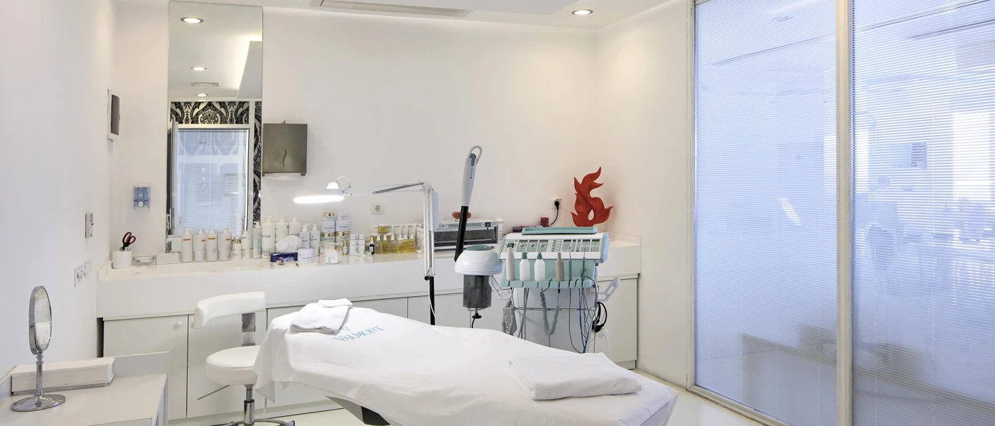 Medical-style treatment room with a machine with wires, unit filled with bottles, a bed covered in white towels and gleaming white walls and flooring