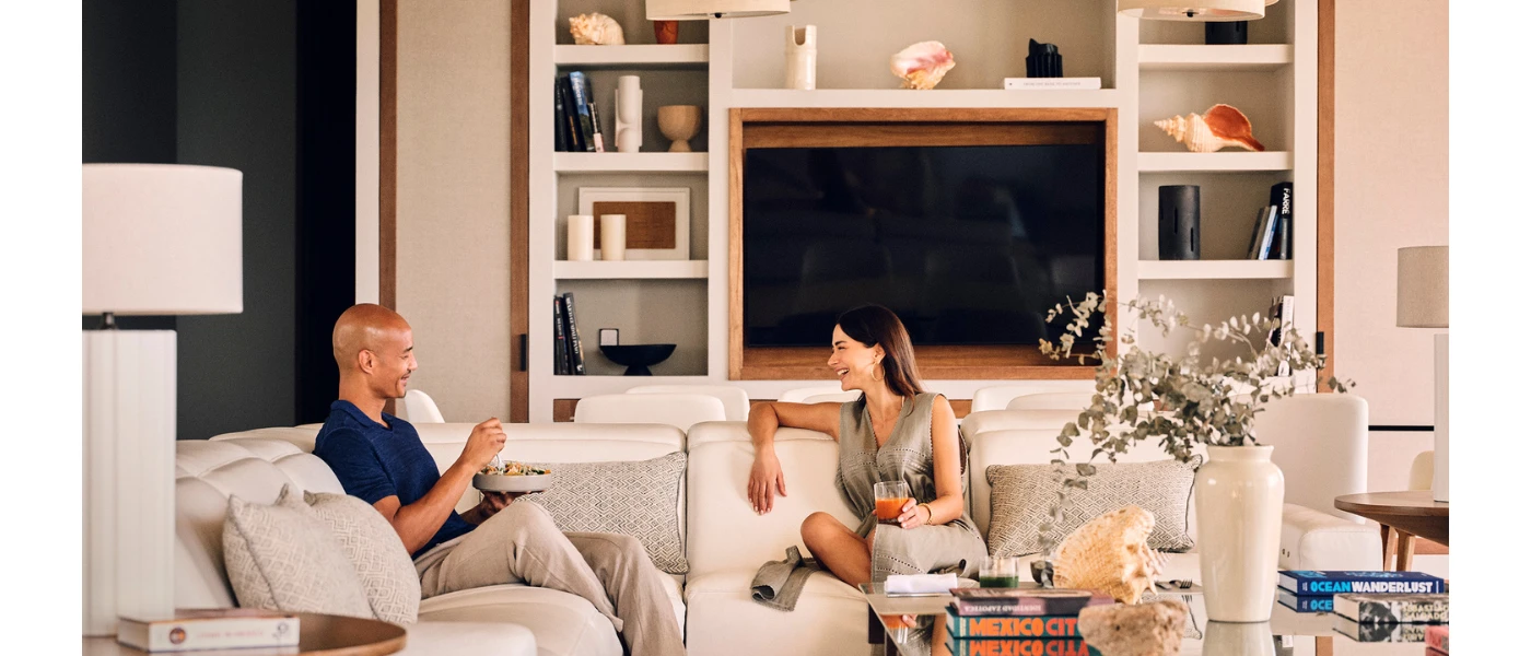 A smiling couple relax on a beige sofa in front of a TV, one with a glass of juice another a bowl of food