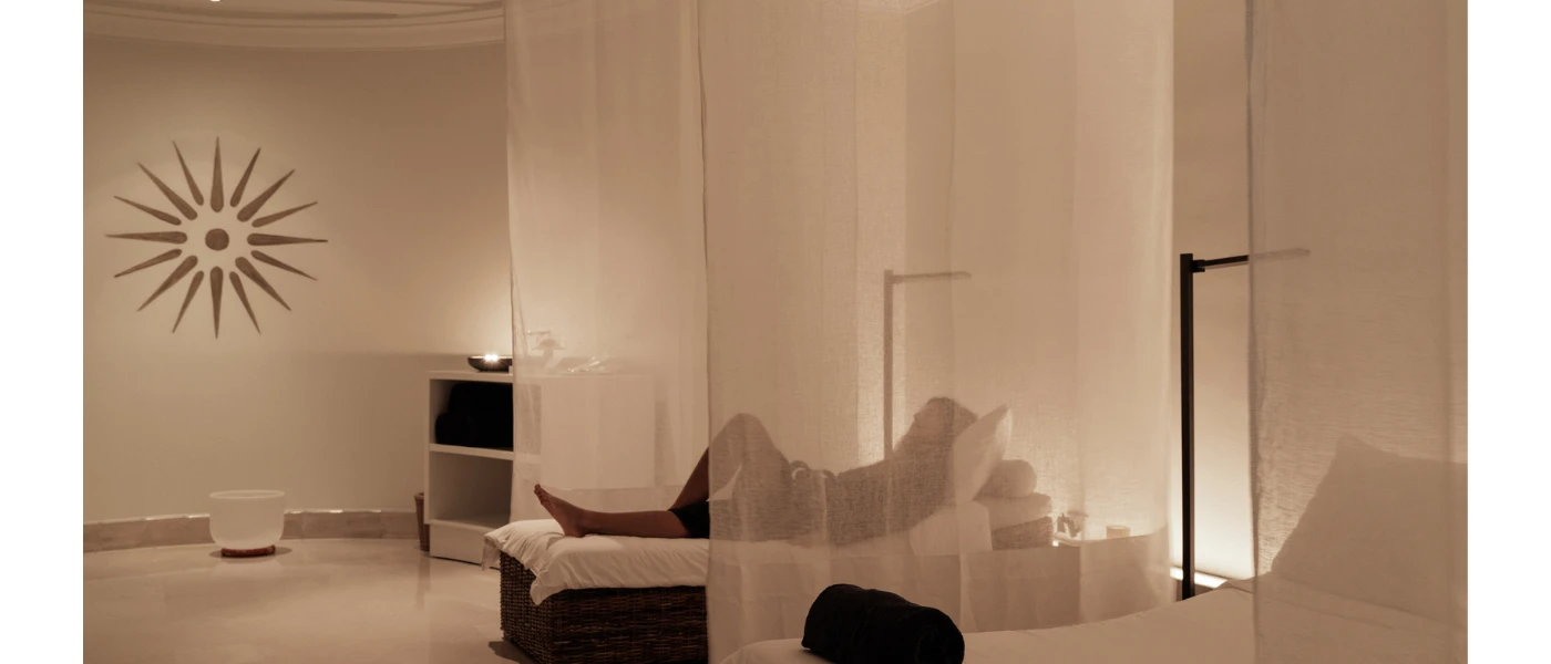 Relaxing softly lift room with white walls, sheer white curtains, candlelight and beds, with a person laid back enjoying the calm