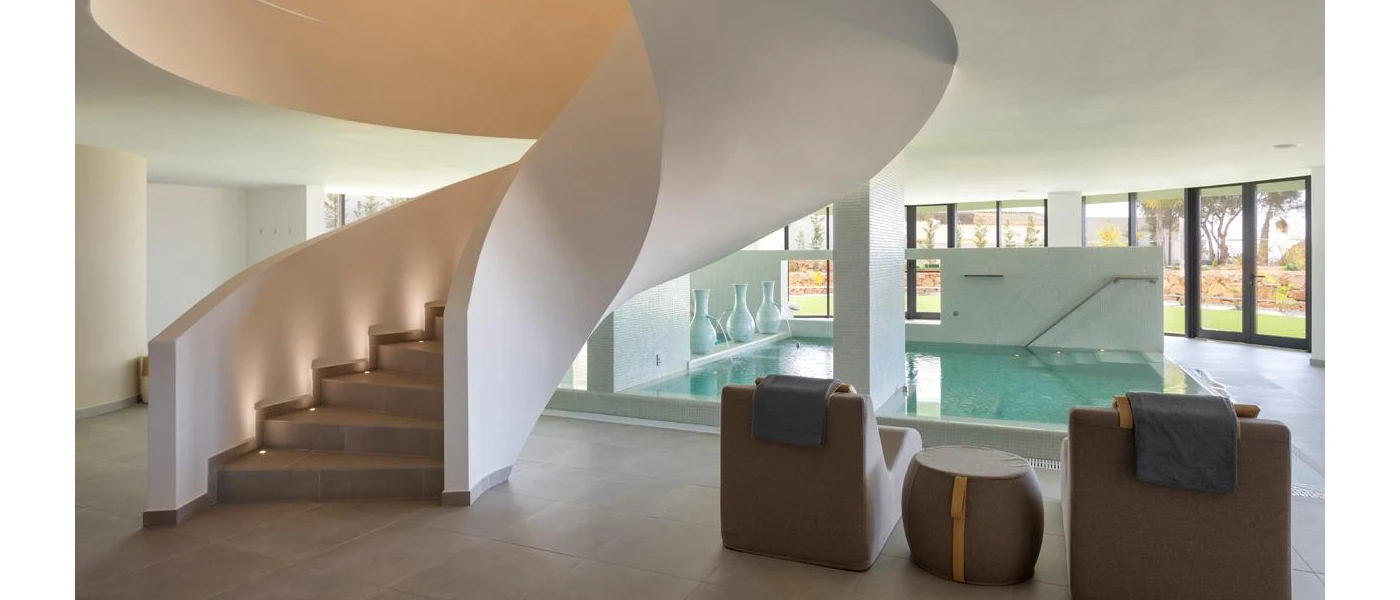 Square indoor pool in a bright room with full-length windows, with white vases and cushioned daybeds lined up at the edges, and a winding staircase leading down to it