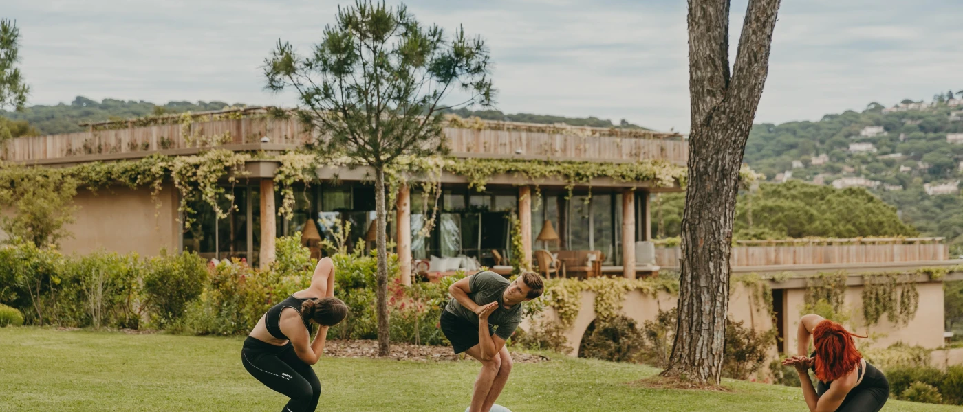 Three people in twisted positions with hands clasped in prayer, on yoga mats in a leafy garden