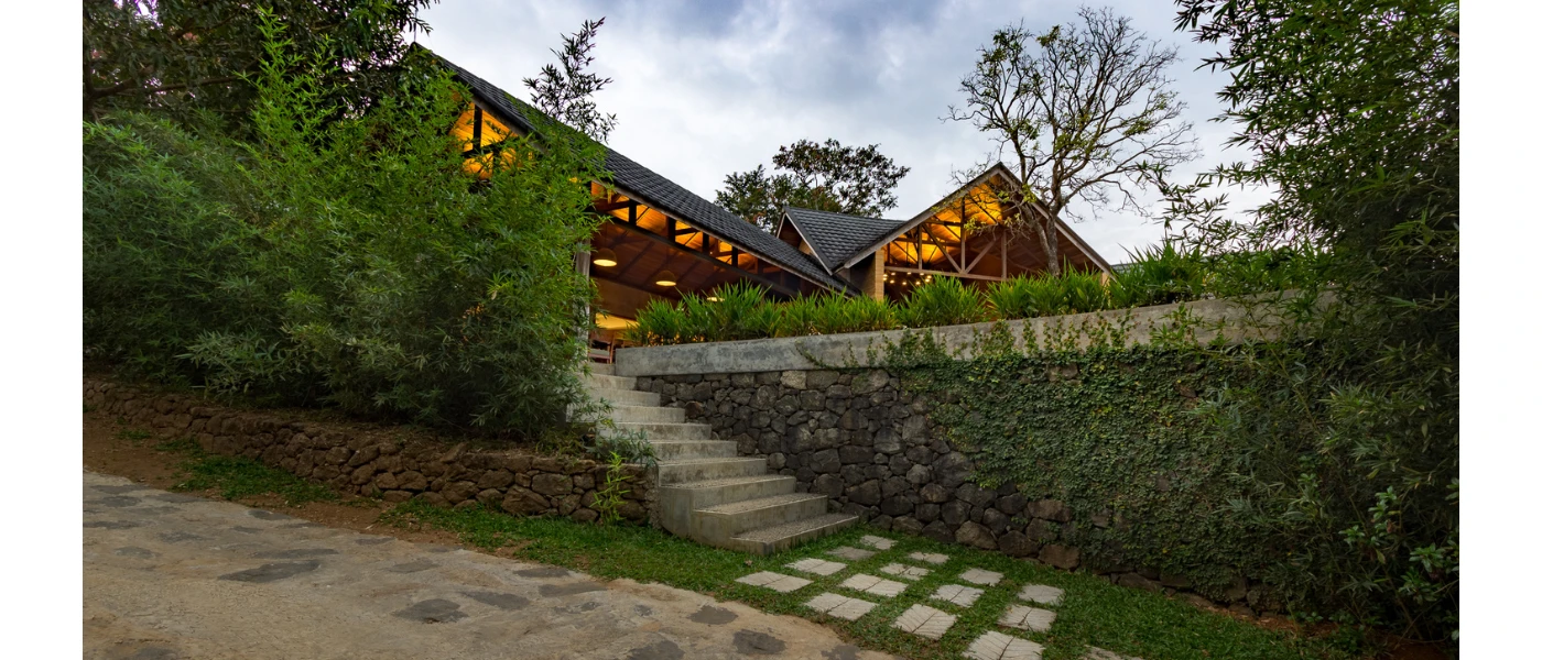 Stone pathways leading to a garden area and a building lit up at twilight, with stone and wood exteriors and a triangular roof