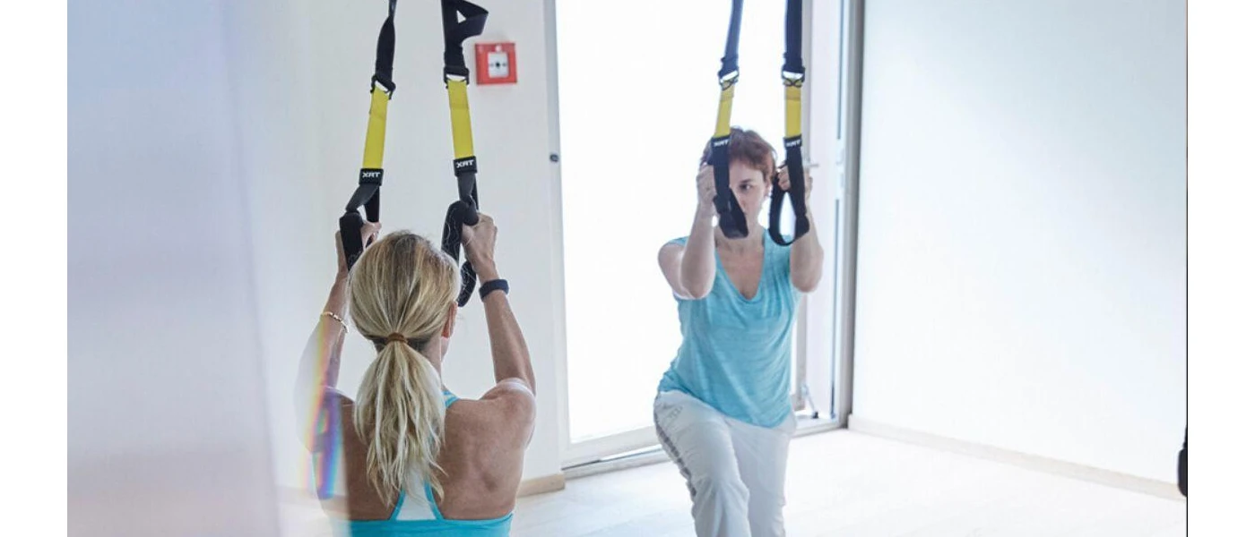 Two women in active wear hang from TRX harnesses in a white studio