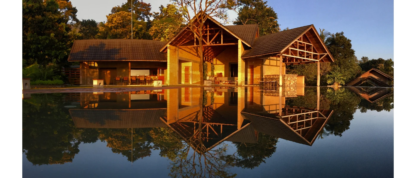 Lake-like swimming pool under a blue sky among tropical greenery, overlooked by a wooden building with triangular rooftops and stilts