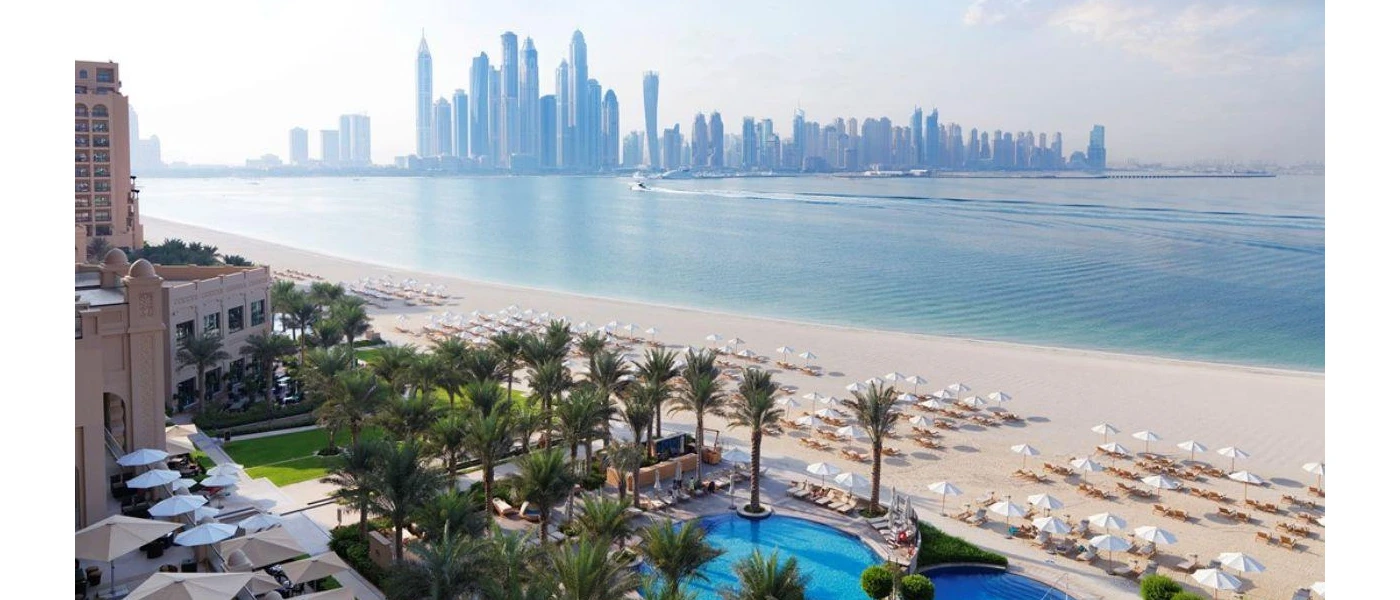 Palm-lined pool and sandy beach looking across the sea to the Dubai skyline