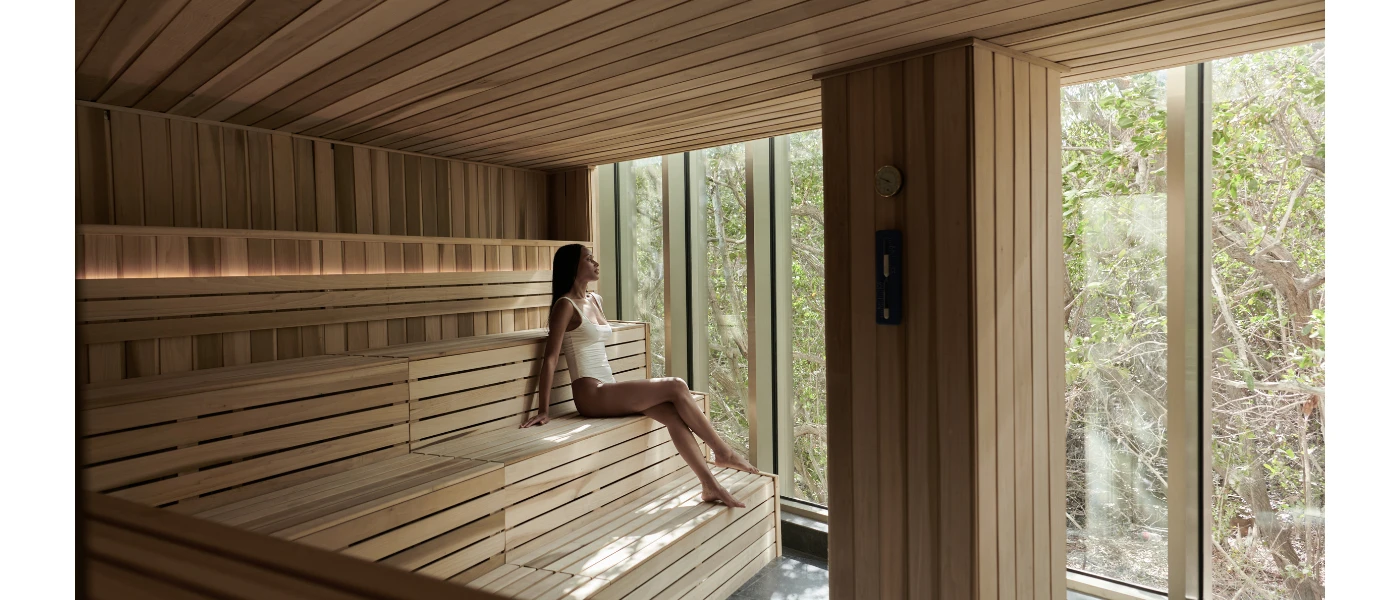 Woman in a white swimsuit sits in a wooden sauna looking out a full-length window at tropical greenery