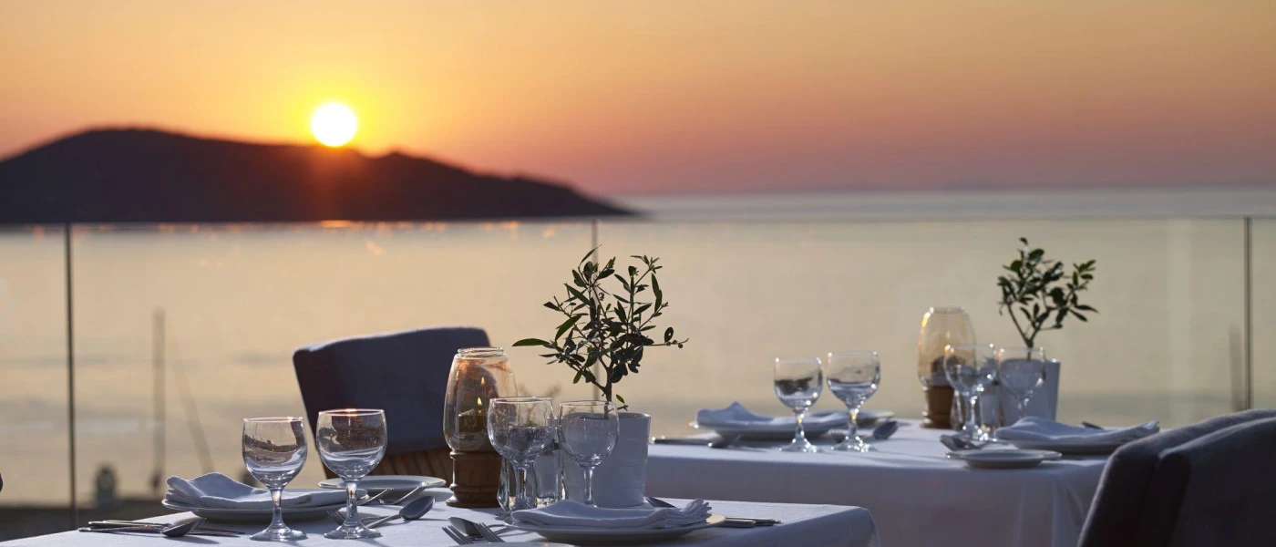 Elegant table set-up with crisp white tablecloths, glassware and olive cuttings on a sea-facing terrace with a glass balcony, as the sun dips behind a hillside opposite