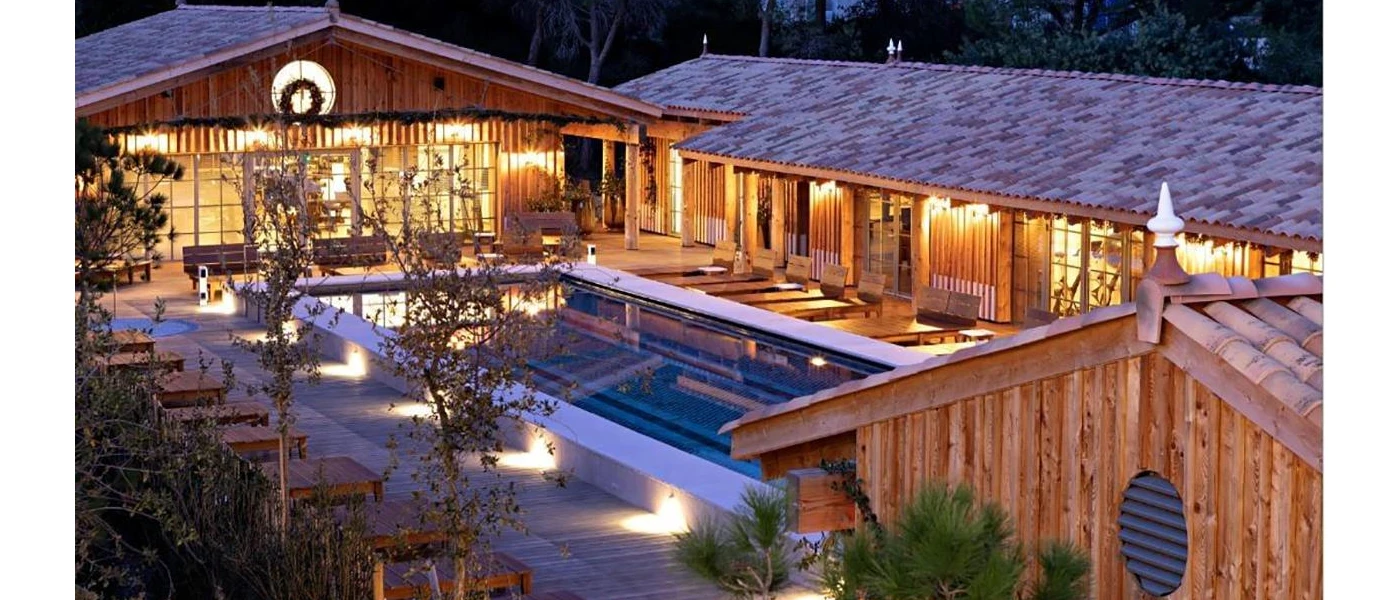 Swimming pool in wooden decking, surrounded by buildings clad in wood, plus wooden loungers, trees and shrubs