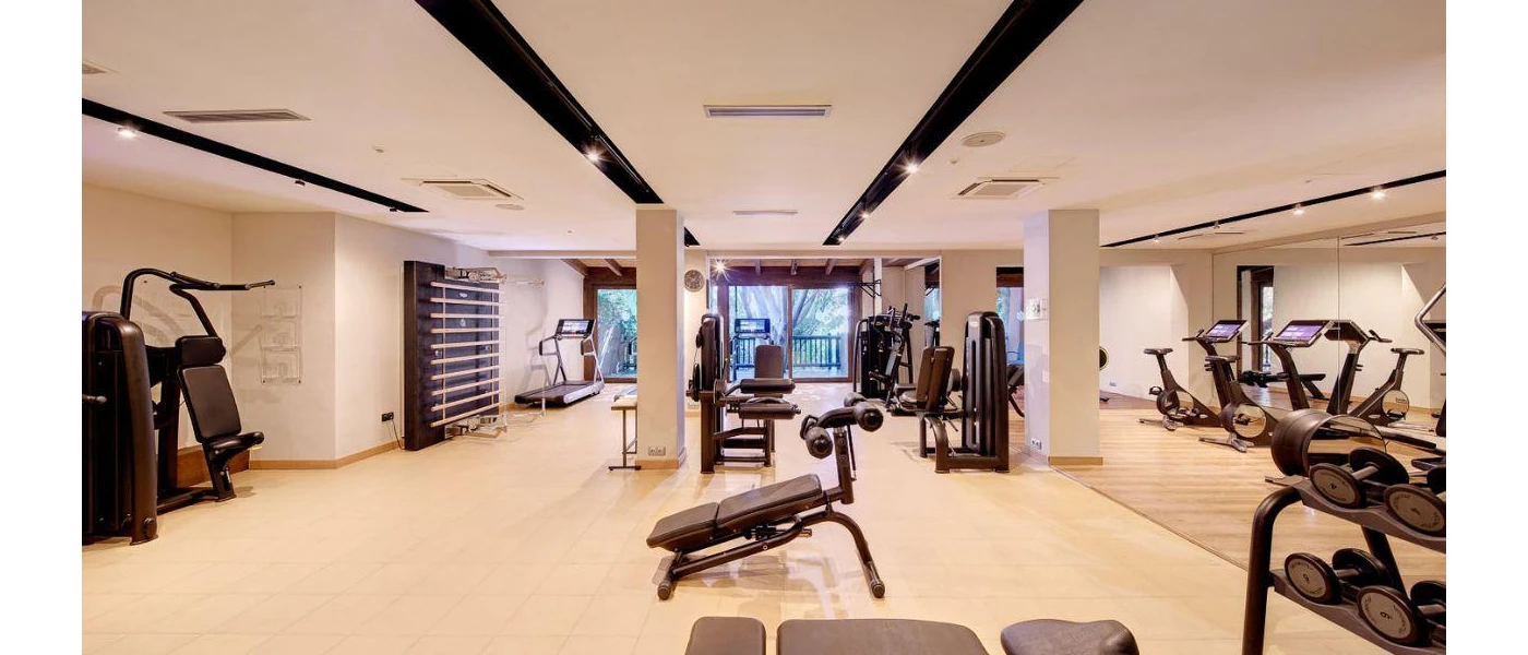 Brightly lit gym with dark beams, bikes, weight machines and benches with a balcony overlooking the gardens