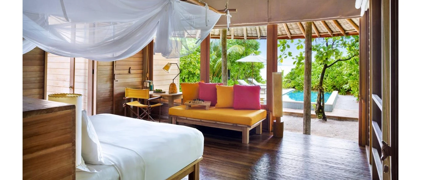 Villa with white bedding with white drapes over the bed, a sofa with pink and yellow cushions, wooden floors and doors open to a swimming pool and greenery beyond