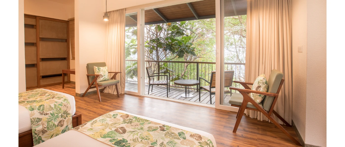 Hotel room with wooden floors and wardrobes, white walls and bedding, green soft furnishings and patio doors open onto a furnished balcony