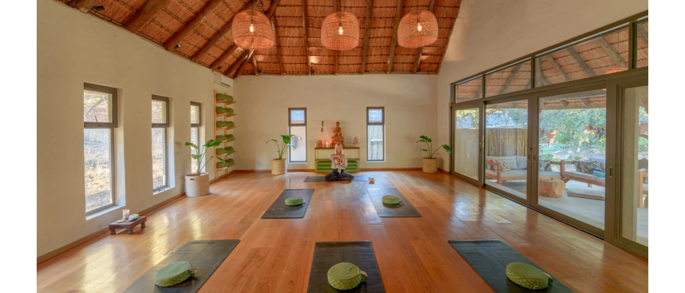 Airy studio with rattan lighting, a wood-beamed ceiling and floor-length windows set up with yoga mats and cushions ready for a class