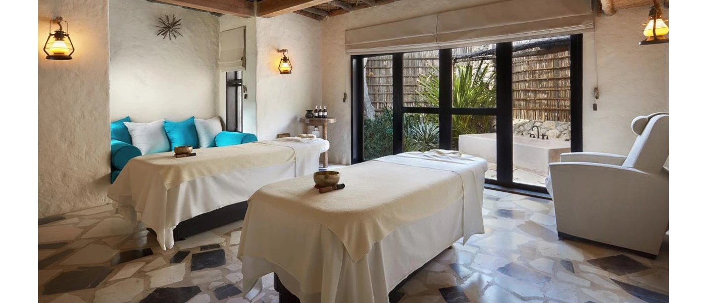 Stone-walled treatment room with two massage beds, both adorned with sound bowls, and full-length windows opening onto a courtyard with a bathtub and tropical greenery