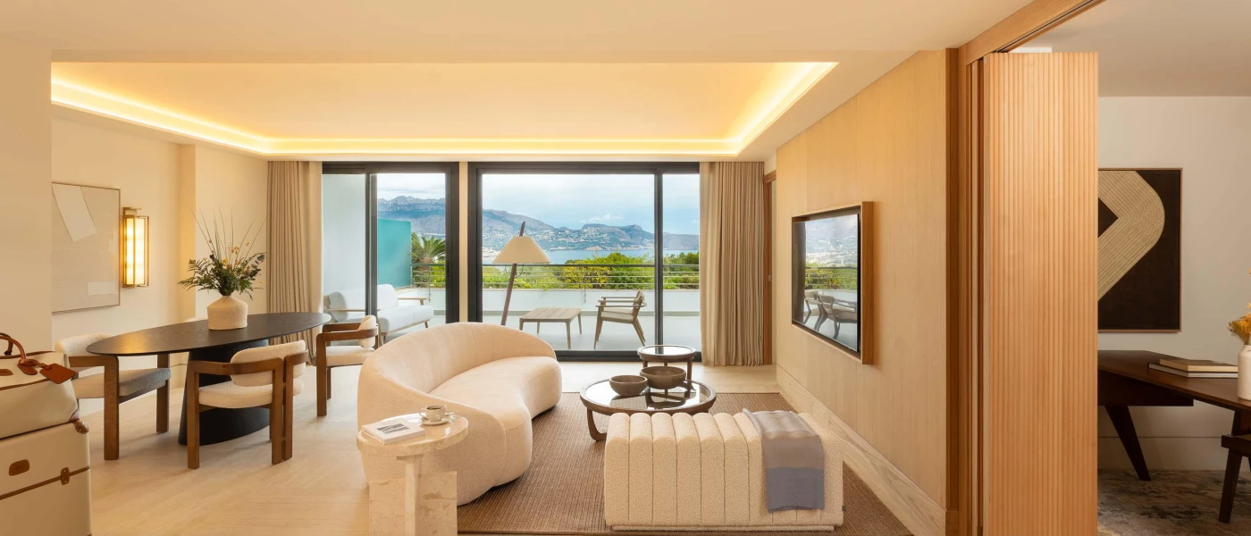 Neutral hotel living area with a cream sofa and wall-hung TV, wooden dining table and floor-to-ceiling balcony doors showcasing furniture and mountain views beyond