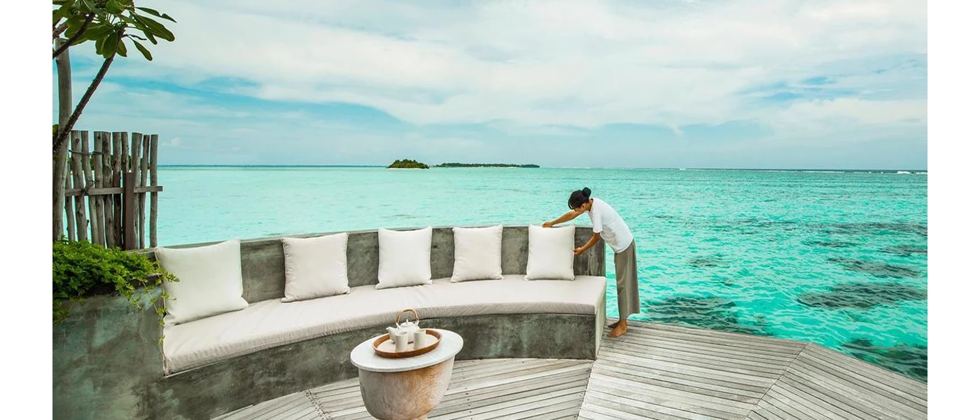 Staff member adjusts an ivory cushion on a cushioned stone sofa, on a wooden deck with direct access to the turquoise ocean