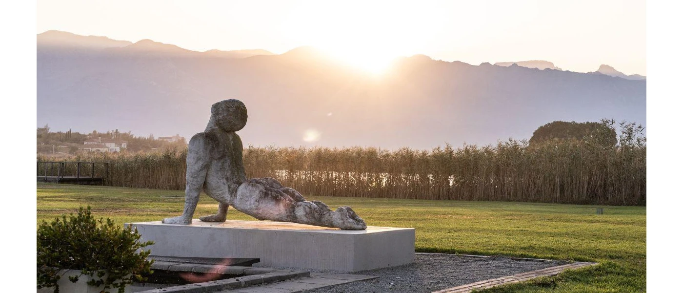 Grey statue of a man in a yoga pose, perched on a concrete block in a grassy lawn