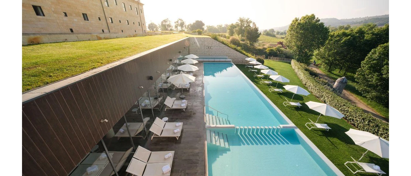 Long, rectangular swimming pool surrounded by white daybeds, loungers and umbrellas, next to grassy lawns and overlooking the countryside