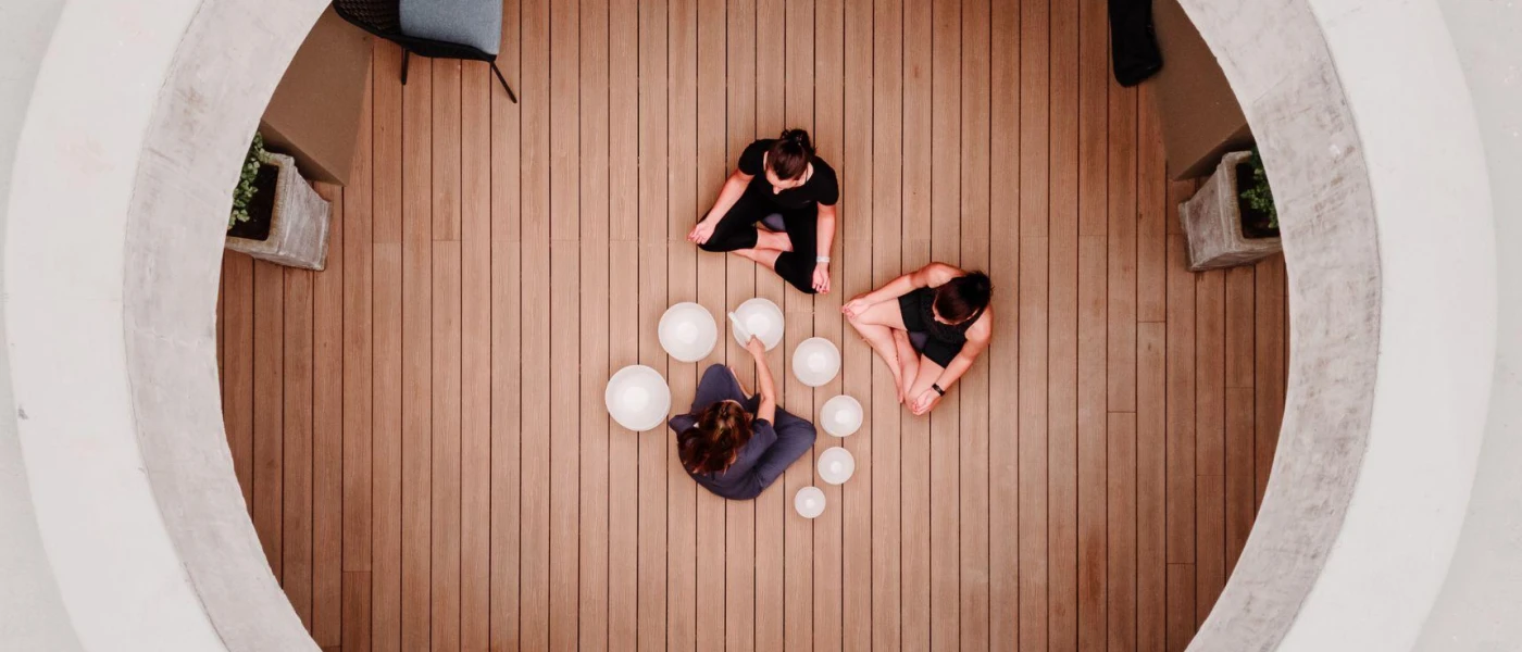 Two women in black sit cross legged in a wood-floored studio while a professional surrounded by bowls carries out sound healing