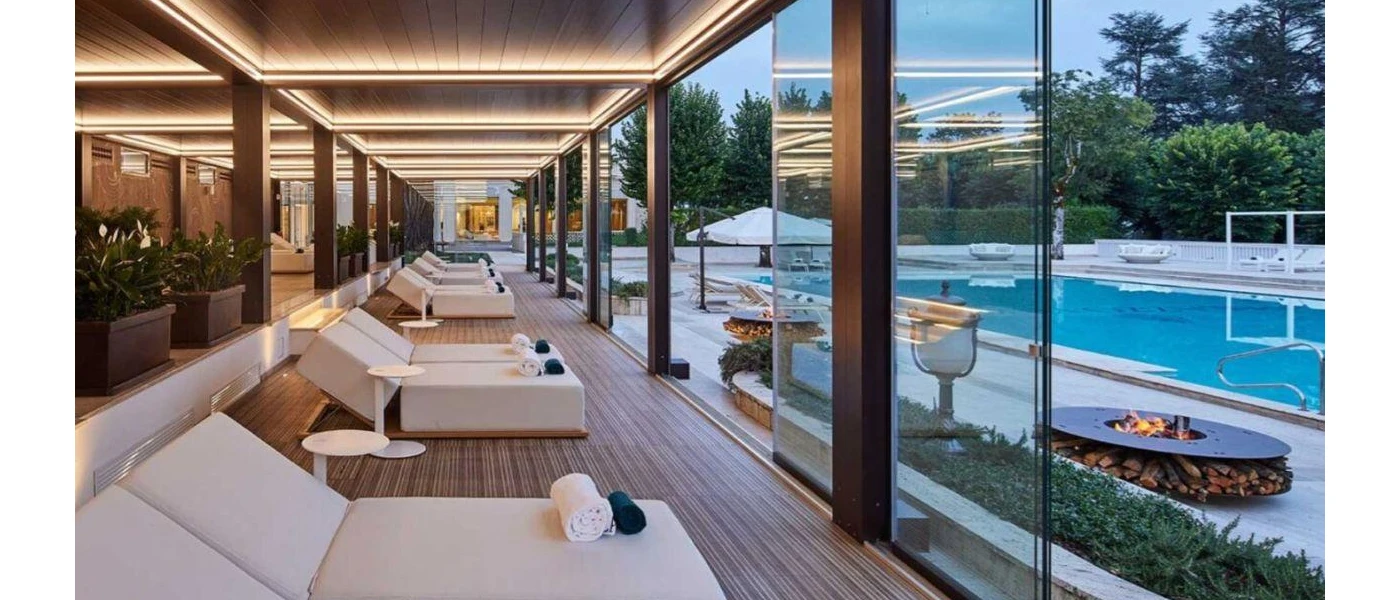 Wooden decking with white daybeds and glass windows overlooking firepits and a swimming pool