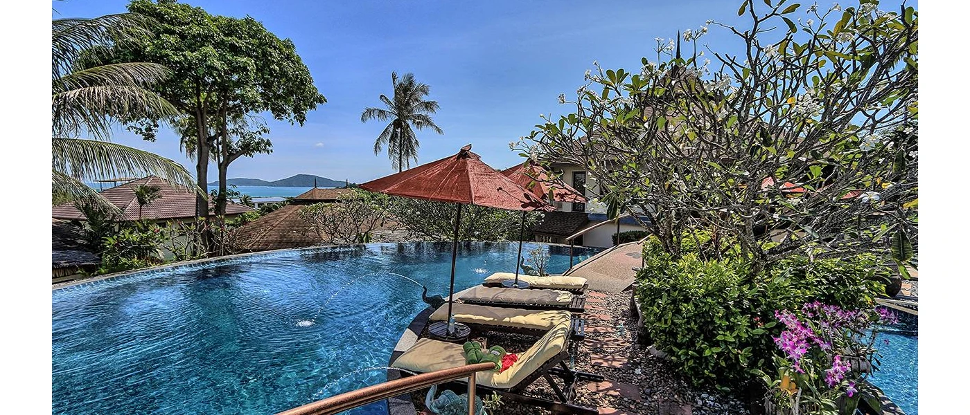 Tranquil pool area surrounded by shrubs and flowers, with a view of the ocean and tiled rooftops - orange umbrellas and loungers line up along the side, and two elephant head fountains spout water into the pool