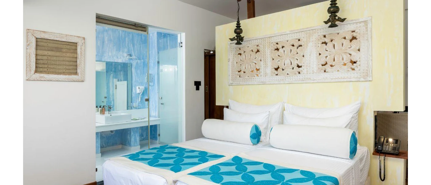 Traditional Sri Lankan room with white bedding, a blue throw and cushions, a headboard with an intricate carved design, and a doorway showing a blue bathroom beyond