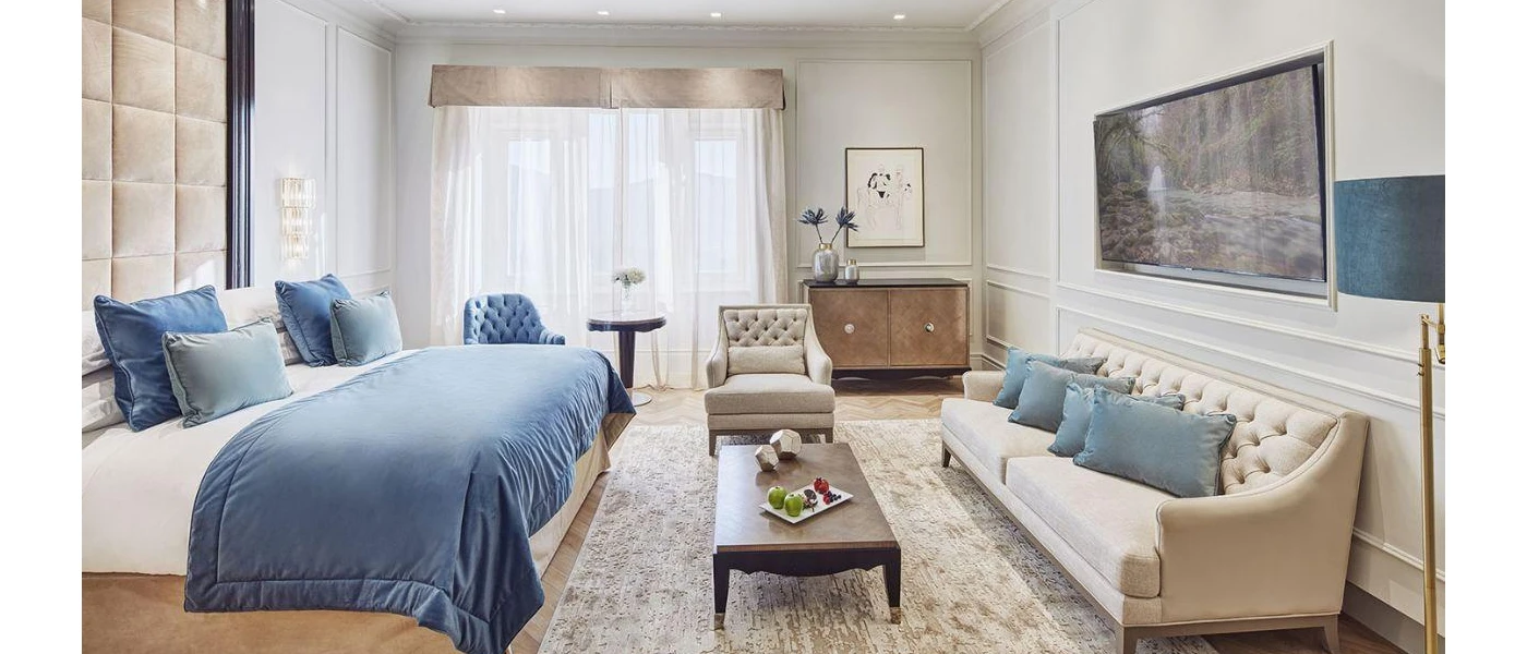 Hotel bedroom with blue cushions and a throw, a beige sofa, wooden coffee table and padded beige velvet headboard