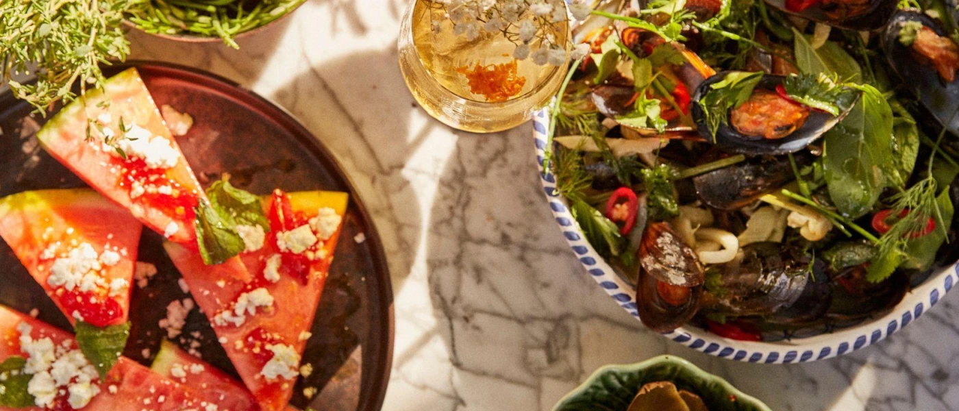 Marble table laid with a plate of watermelon sprinkled with feta cheese, and a bowl of green salad with mussels