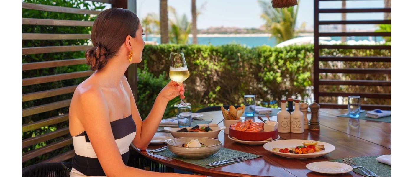 Woman in striped strapless top and gold earrings sips white wine with a sea view, and a table filled with seafood