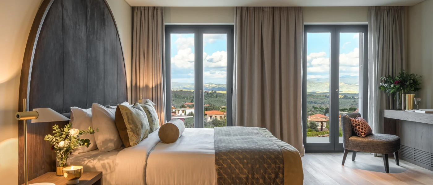 Hotel room with a stylish curved velvet headboard, white bedding with neutral cushions, floor-to-ceiling windows with neutral curtains and a desk area with a gold vase and flowers