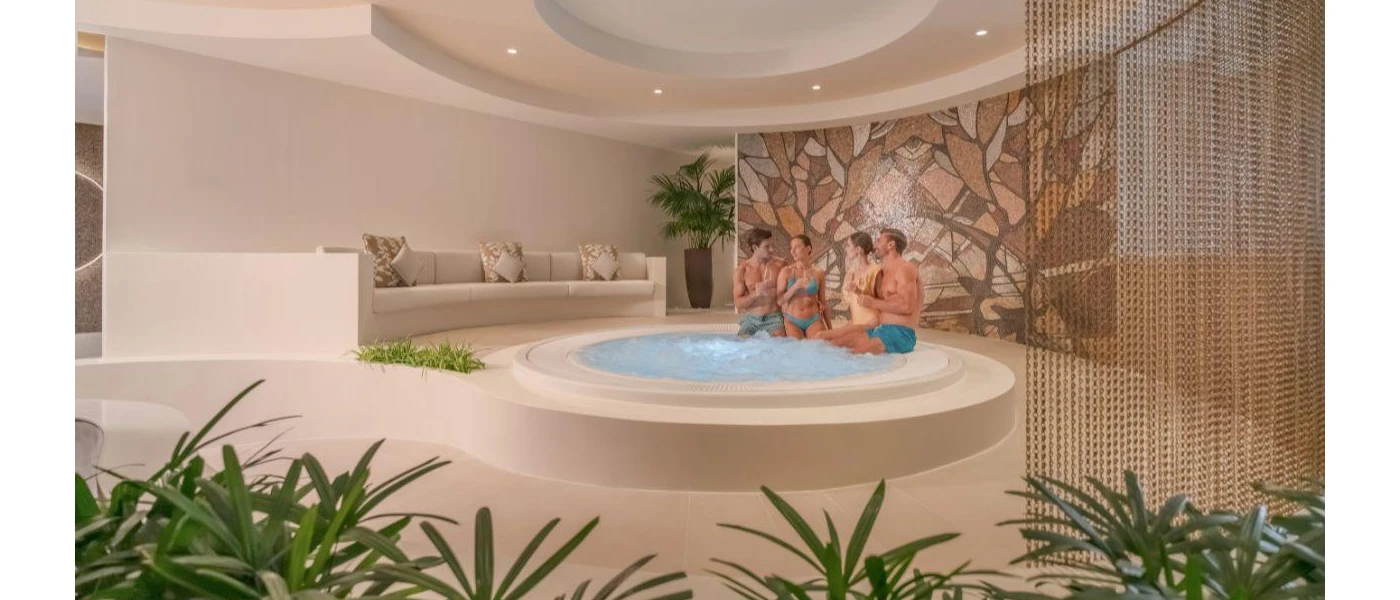 Two couples sit on the edge of a whirlpool bath next to a neutral sofa and palms with a mosaic tiled wall behind them