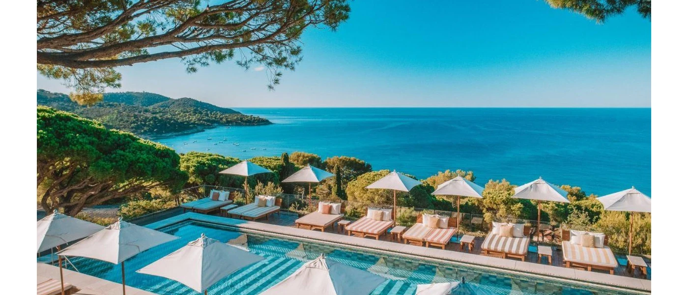 Elevated swimming pool with a sea view, lined with pink and white striped loungers, white umbrellas and greenery